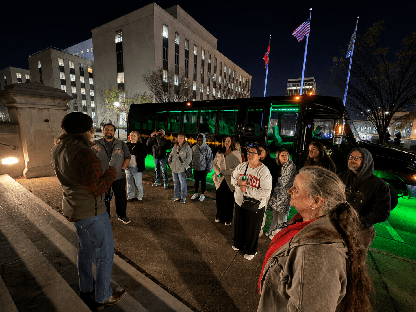 Group of people listening to a guide near a lit bus at night, with buildings and flags in the background.