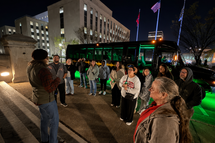 Group of people listening to a guide near a lit bus at night, with buildings and flags in the background.