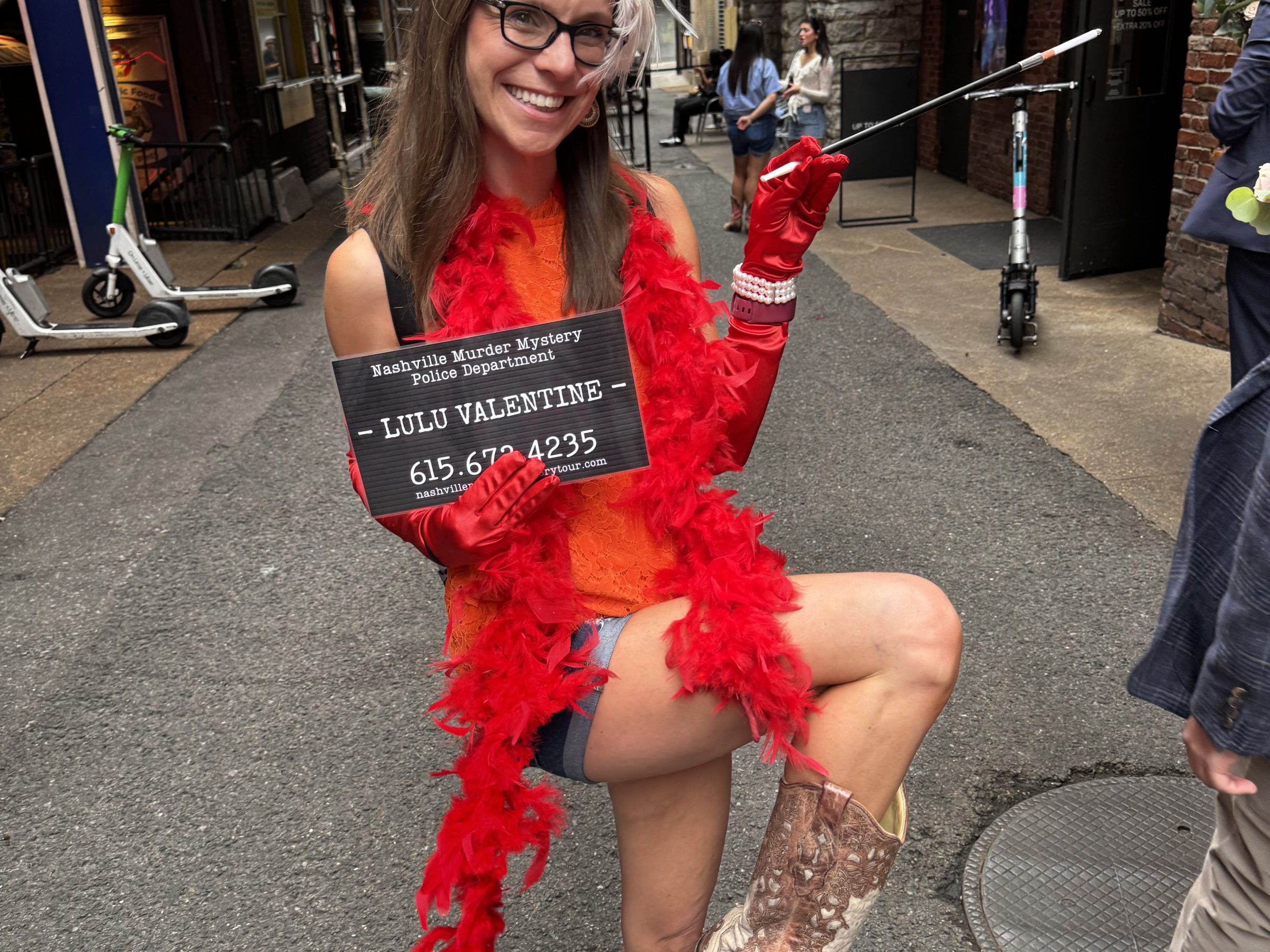 Nashville murder mystery pub crawl 2 Woman in red boa, feather headband, holding sign and cane, wearing cowboy boots in city street.