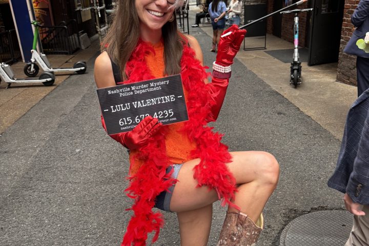 Nashville murder mystery pub crawl 2 Woman in red boa, feather headband, holding sign and cane, wearing cowboy boots in city street.