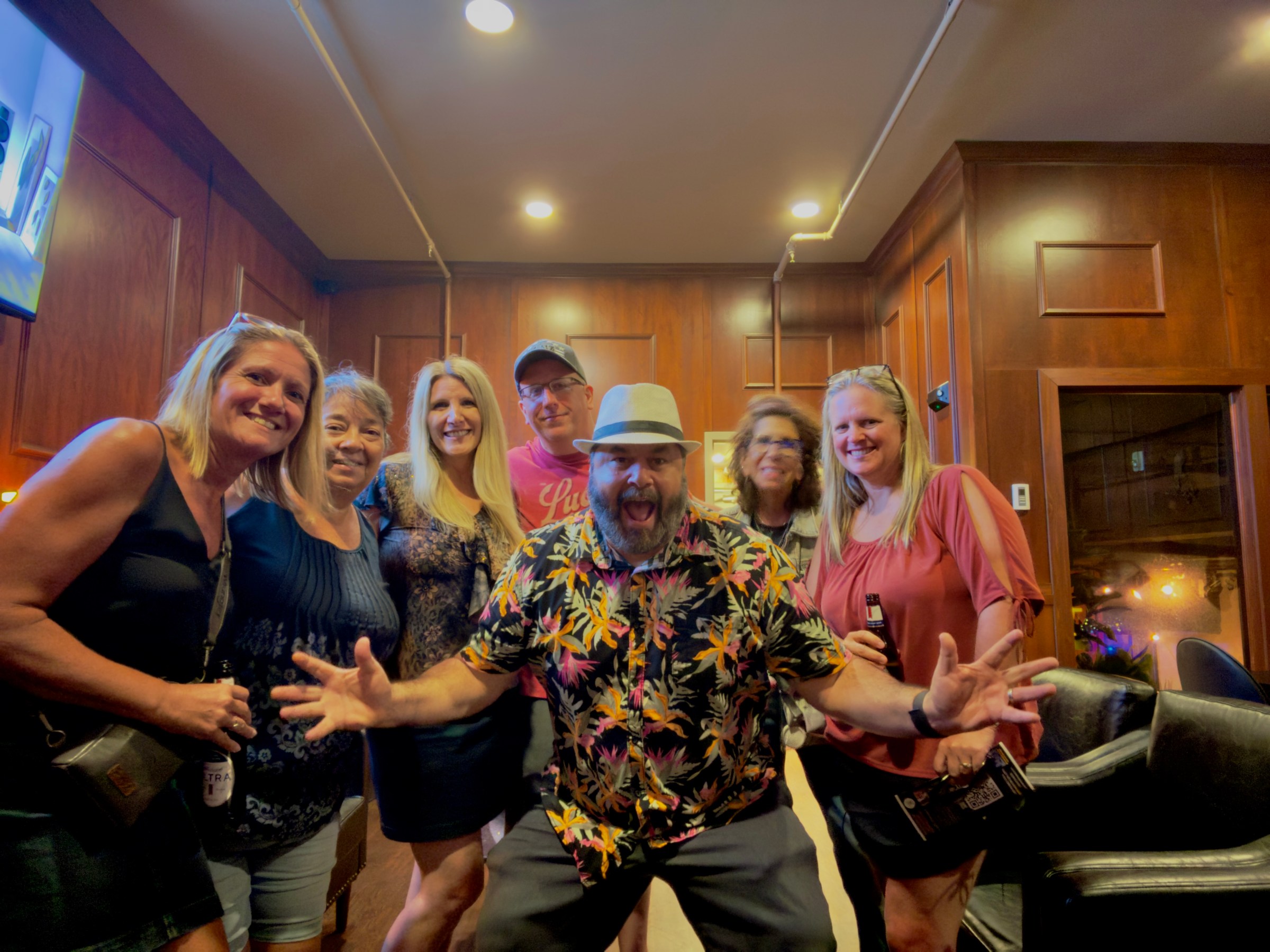 Seven smiling adults posing in a wood-paneled room, one man in a floral shirt is making a playful gesture. murder mystery guest detectives in nashville