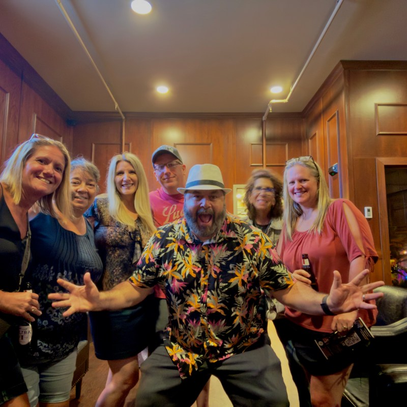 Seven smiling adults posing in a wood-paneled room, one man in a floral shirt is making a playful gesture. murder mystery guest detectives in nashville