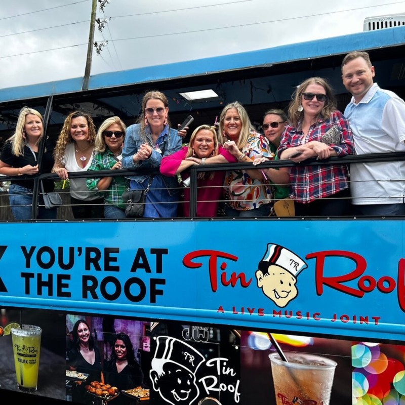 Group of people smiling on a blue bus with 'Tin Roof' signage.