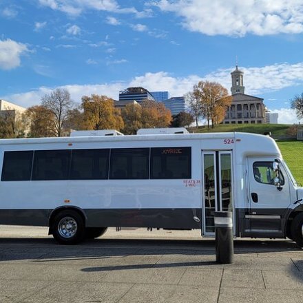 White shuttle bus parked on city street with buildings and trees in background.
