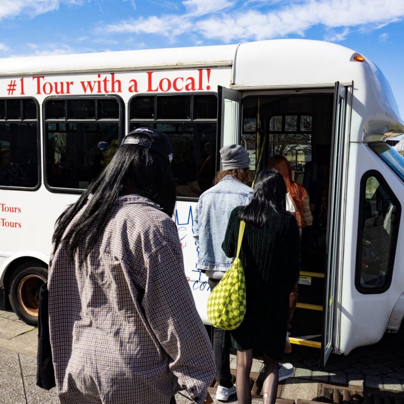 Group of people boarding a tour bus labeled '#1 Tour with a Local!' on a sunny day.