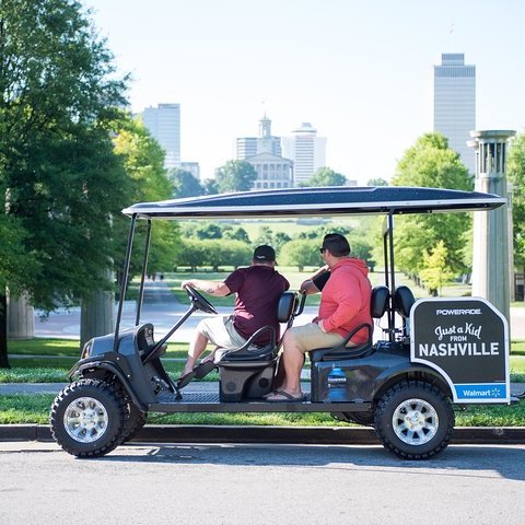 Two people ride a golf cart branded with 'Nashville' in a park, city skyline visible in the background.