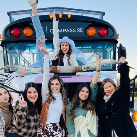 Six women posing joyfully in front of a bus with steer horns on its grill.