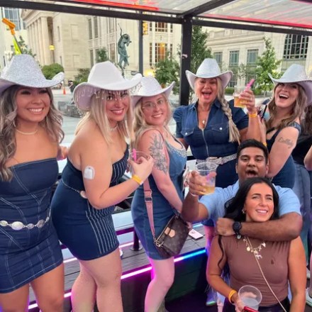 Group of people in cowboy hats and denim outfits smiling and holding drinks on a party bus.