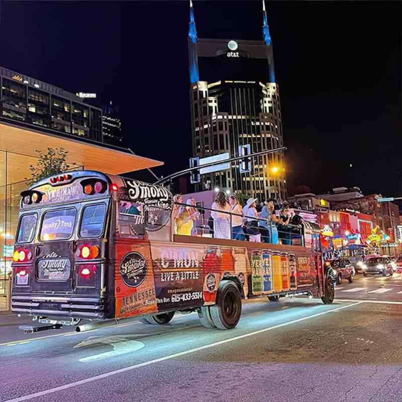 Open-air party bus with people in a city at night, tall building and neon lights in the background.