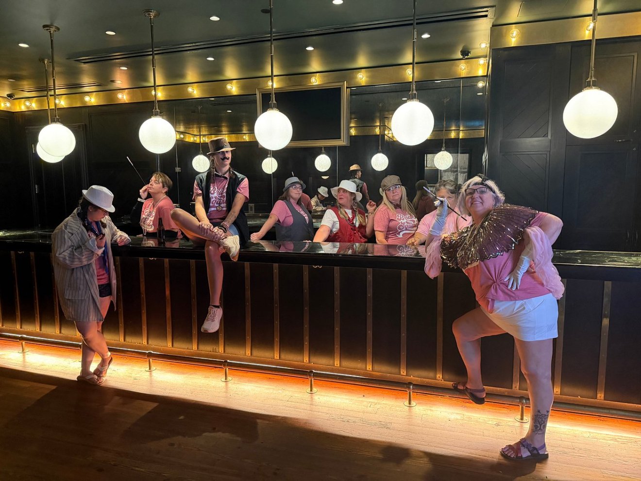 People in pink and white outfits posing playfully at a bar with hanging globe lights.